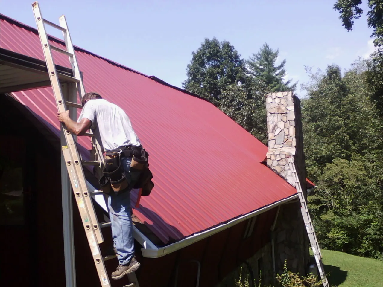 Roofer on a ladder beside a red metal roof and stone chimney — Soffit & Fascia Repair by Roofing Brothers in Hayfork